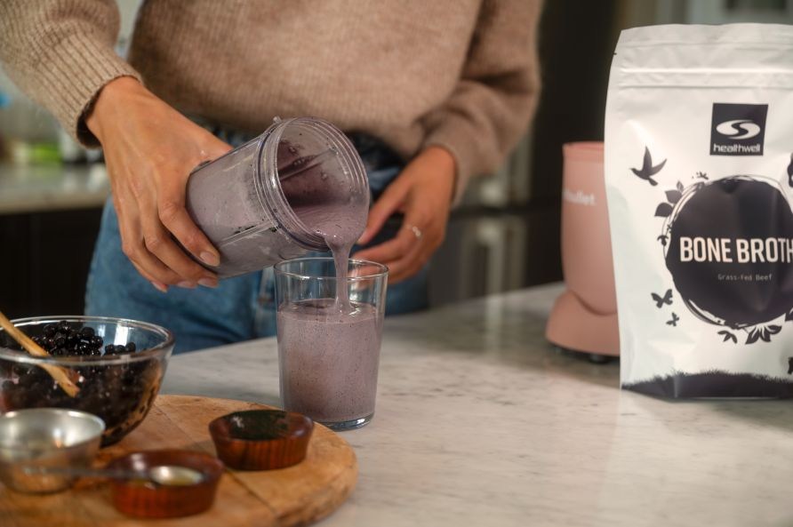 En kvinna h�ller upp bl�b�rssmoothie med benbuljong fr�n en mixer i ett glad. P� bordet syns en sk�l med bl�b�r och en p�se Healthwell Benbuljong EKO.