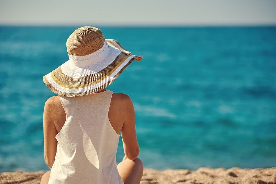 Kvinna med solhatt och linne som sitter på en strand  och kollar ut över havet.