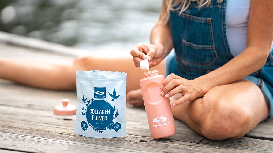 Kvinna sitter p� en brygga och tar en skoppa av Healthwell Collagen Pulver Bovint och h�ller i en rosa vattenflaska.