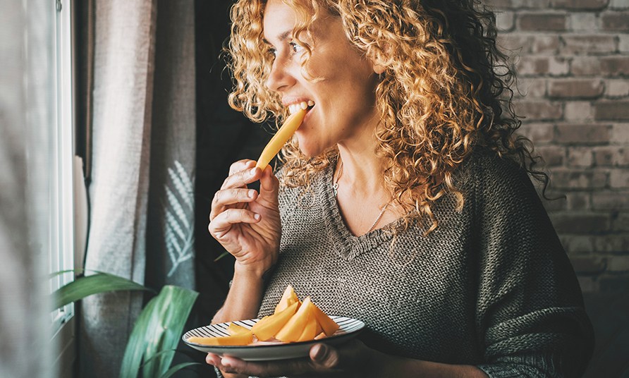 En kvinna str vid ett fnster och ter en bit frukt.
