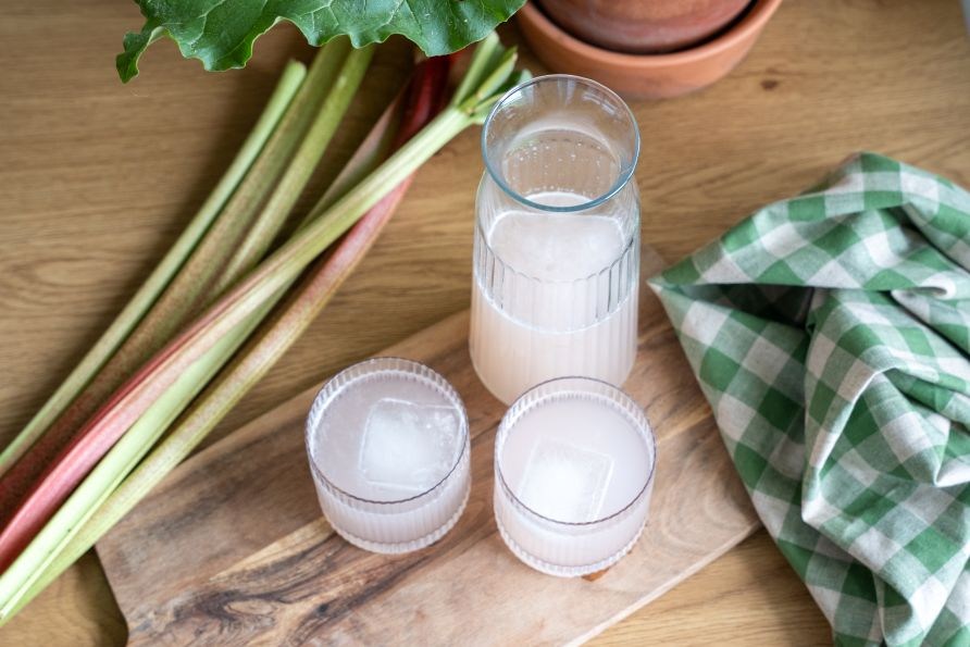 Hemgjord rabarbersaft med isbitar serverad i glas och karaff, med f�rska rabarberstj�lkar bredvid p� ett tr�br�de.