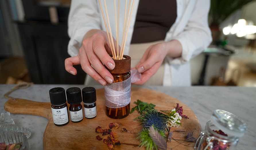 Trbricka med ingredienser fr att blanda din egen rumsdoft och en kvinna som rcker sig efter en glasflaska med trpinnar.