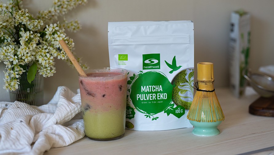 Healthwell Matcha Pulver EKO i originalfrpackning, iced strawberry matcha latte i ett glas med ett bambusugrr, visp fr matchalatte, blombukett i en vas och en kkshandduk p ett bord. 